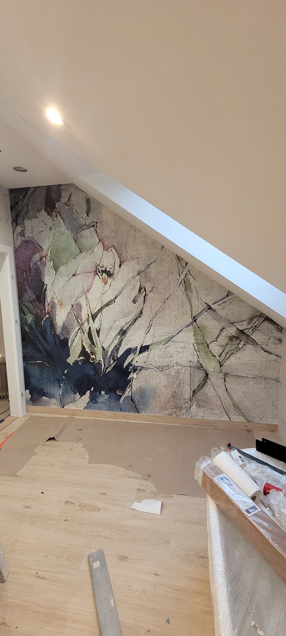 Ściana w pokoju na poddaszu pokryta tapetą z motywem kwiatowym w odcieniach szarości, zieleni i fioletu, podłoga zabezpieczona kartonem, widoczne narzędzia i rolka tapety.