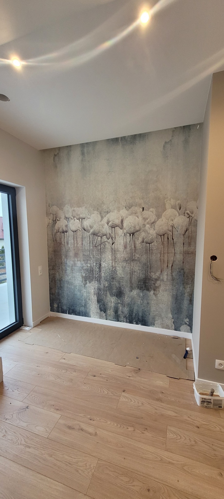 Pokój po remoncie z nową tapetą z motywem flamingów i świeżo ułożoną podłogą z jasnego drewna, zabezpieczoną kartonem podczas prac wykończeniowych.