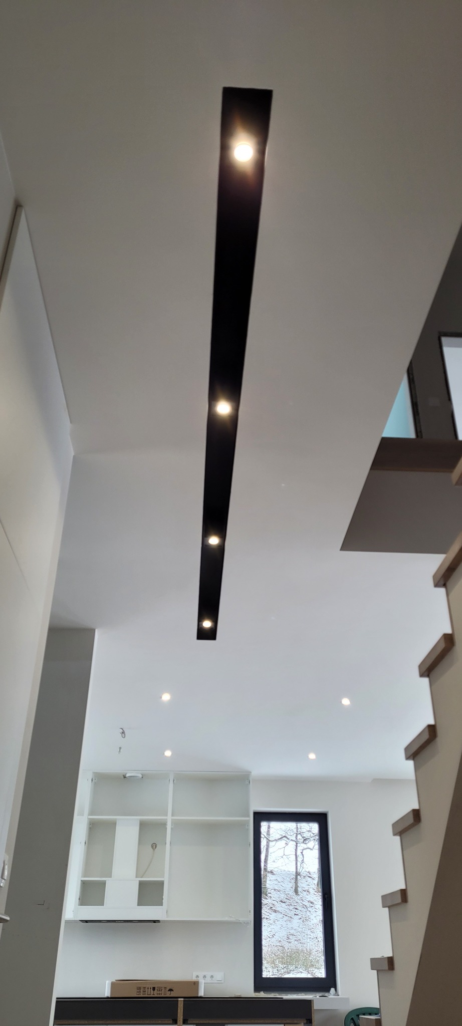 Nowoczesne oświetlenie sufitowe z czarną listwą i punktowymi lampami w jasnym wnętrzu z widoczną częścią kuchni i schodami.