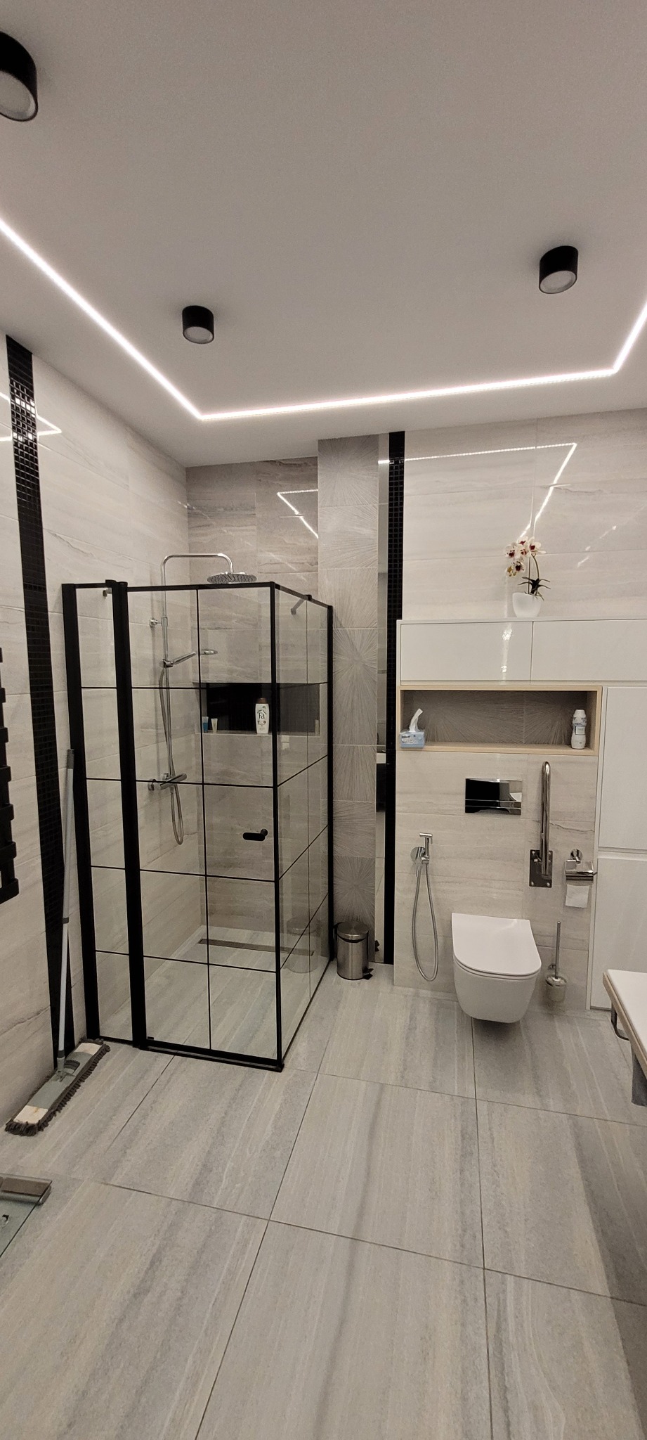 Nowoczesna łazienka z kabiną prysznicową typu walk-in z czarnymi profilami, podwieszaną toaletą, szarą glazurą i oświetleniem LED na suficie.
