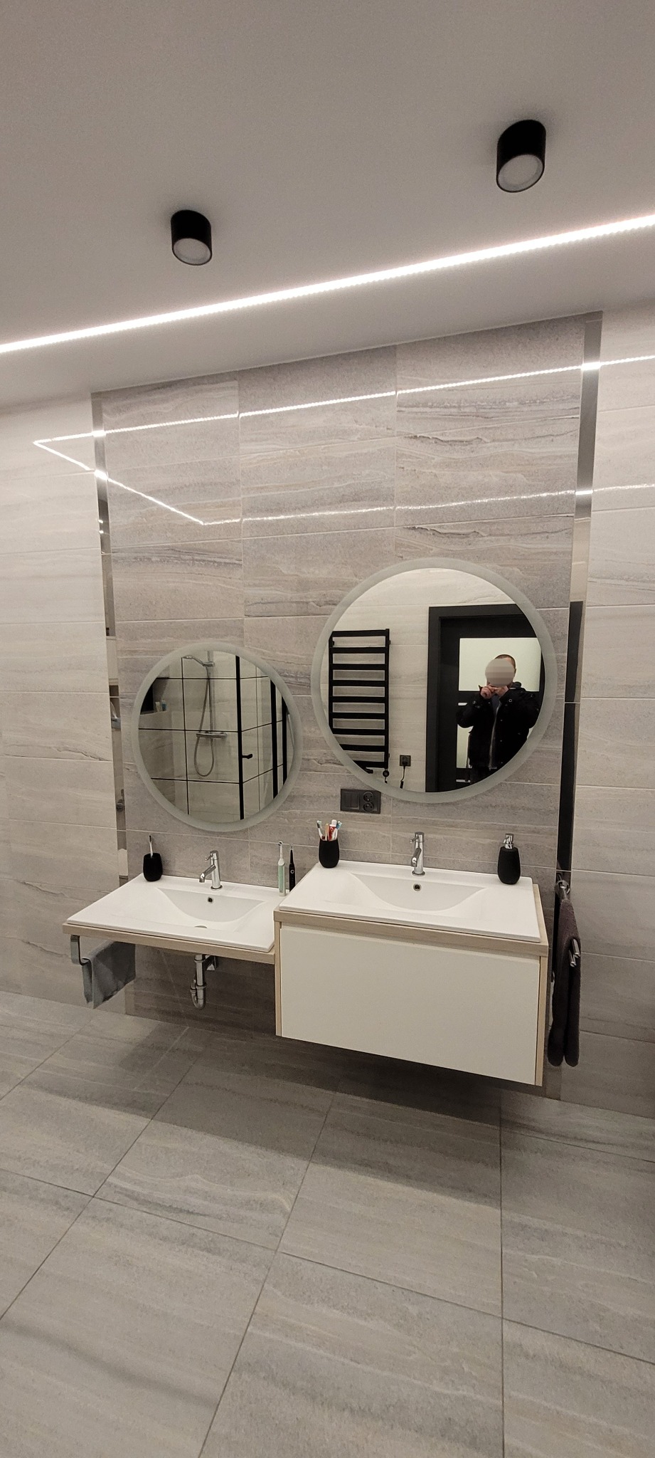 Nowoczesna łazienka z dwoma okrągłymi lustrami z podświetleniem LED, umywalkami nablatowymi i płytkami imitującymi naturalny kamień, z widocznym odbiciem osoby robiącej zdjęcie.