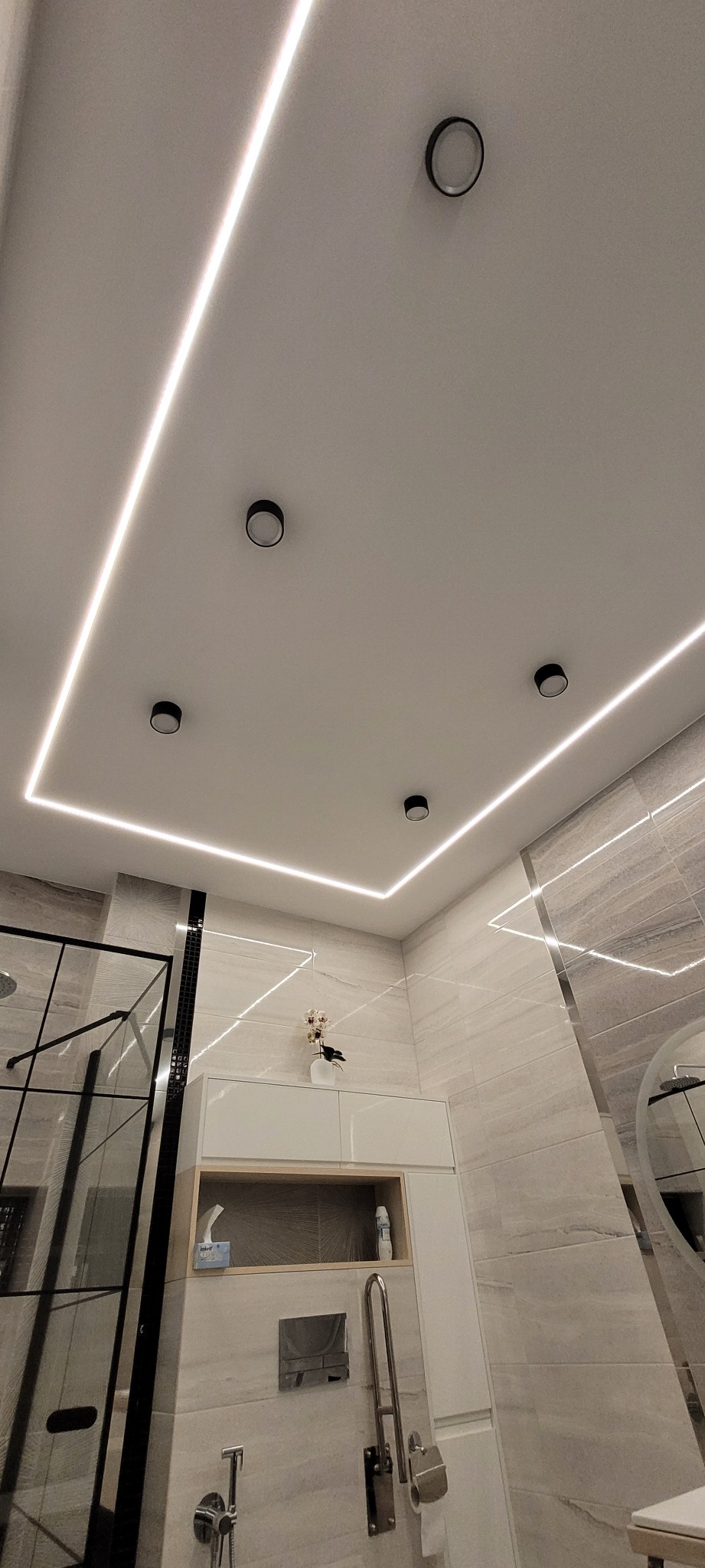 Nowoczesna łazienka z podświetlanym sufitem i czarnymi punktowymi lampami, jasne płytki imitujące marmur, kabina prysznicowa ze szklaną ścianką, biała szafka z półką i dekoracją.