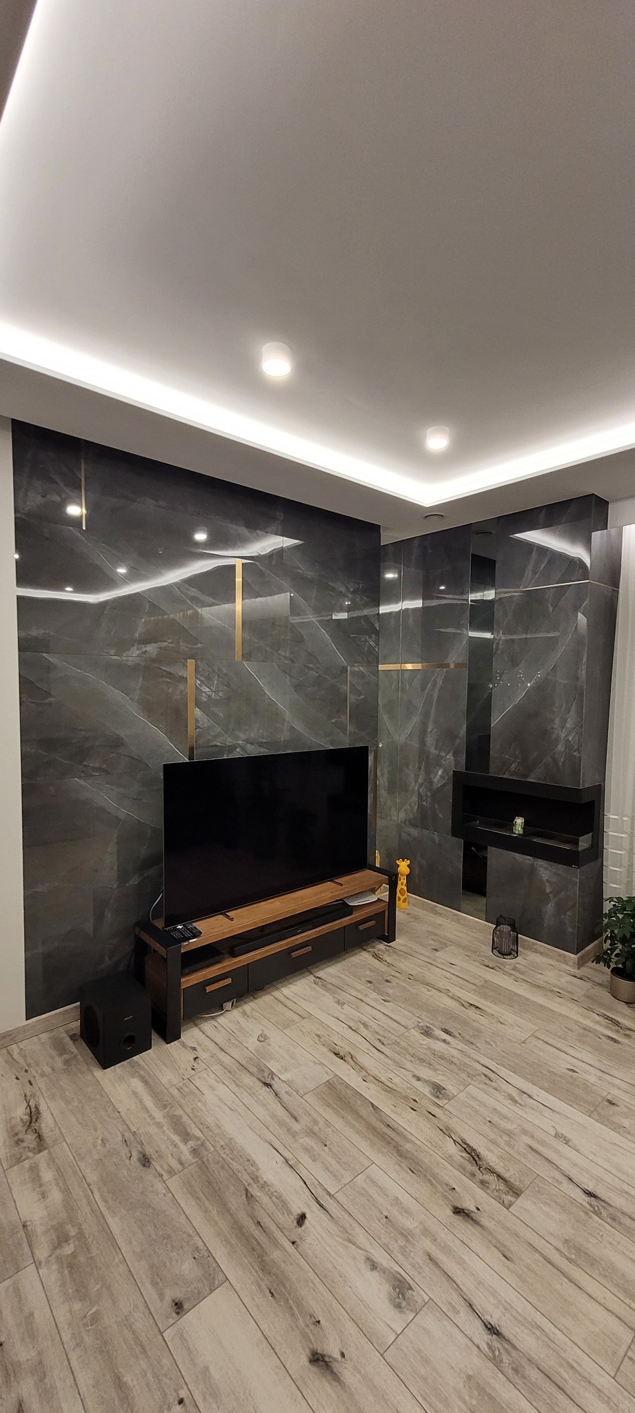Elegancki salon z szarymi płytkami imitującymi marmur na ścianie, podkreślonymi złotymi wstawkami, drewnianą podłogą, telewizorem na szafce RTV i podwieszanym sufitem z oświetleniem LED.