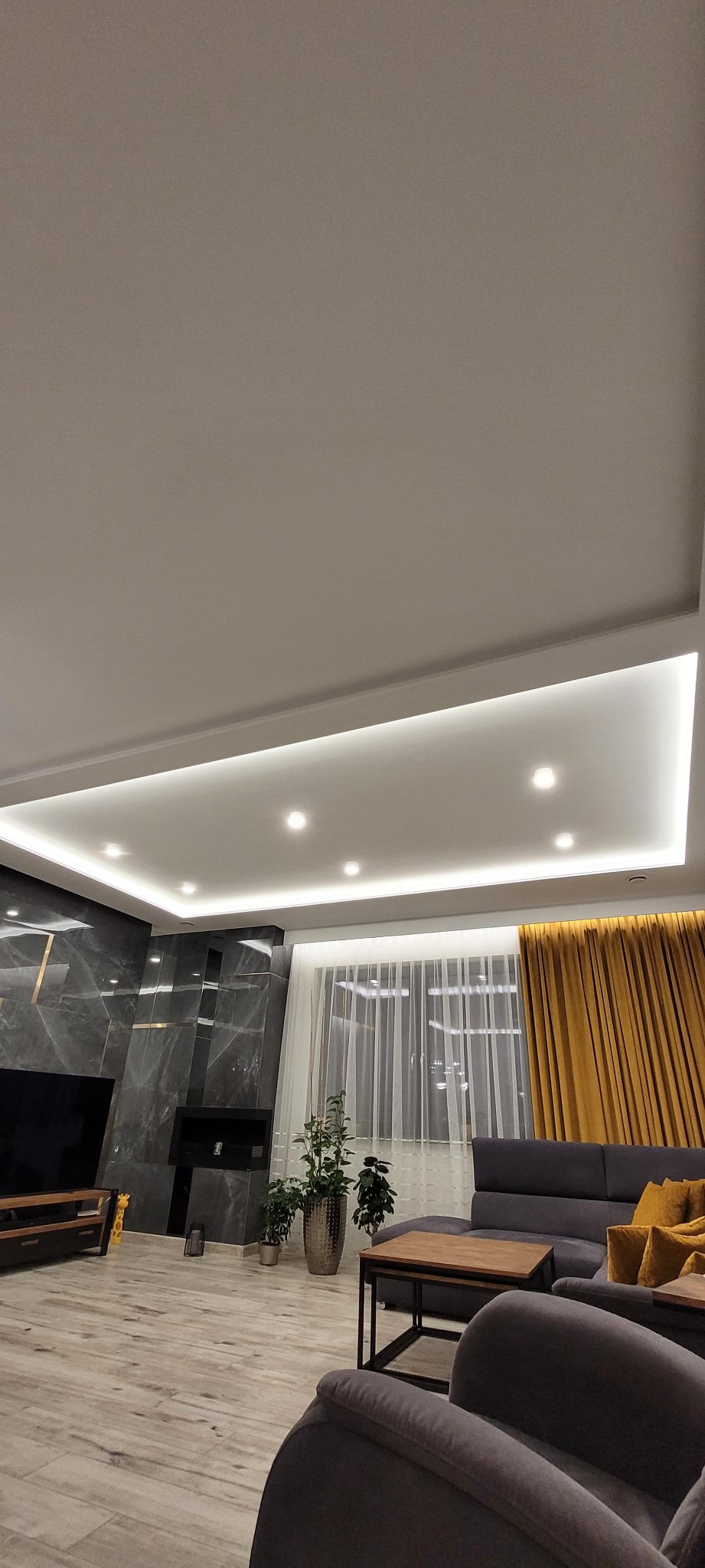 Nowoczesny salon z podwieszanym sufitem z oświetleniem LED, szarą ścianą z imitacją marmuru, telewizorem, drewnianą podłogą i szarą sofą z żółtymi poduszkami.