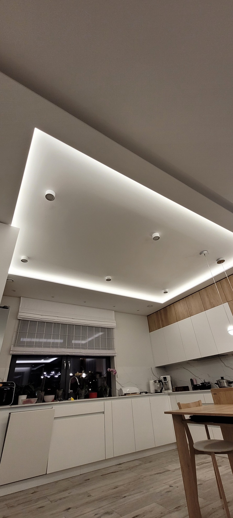 Nowoczesna kuchnia z oświetleniem LED w suficie podwieszanym, widoczne okno z roletami rzymskimi, białe szafki i drewniane akcenty.