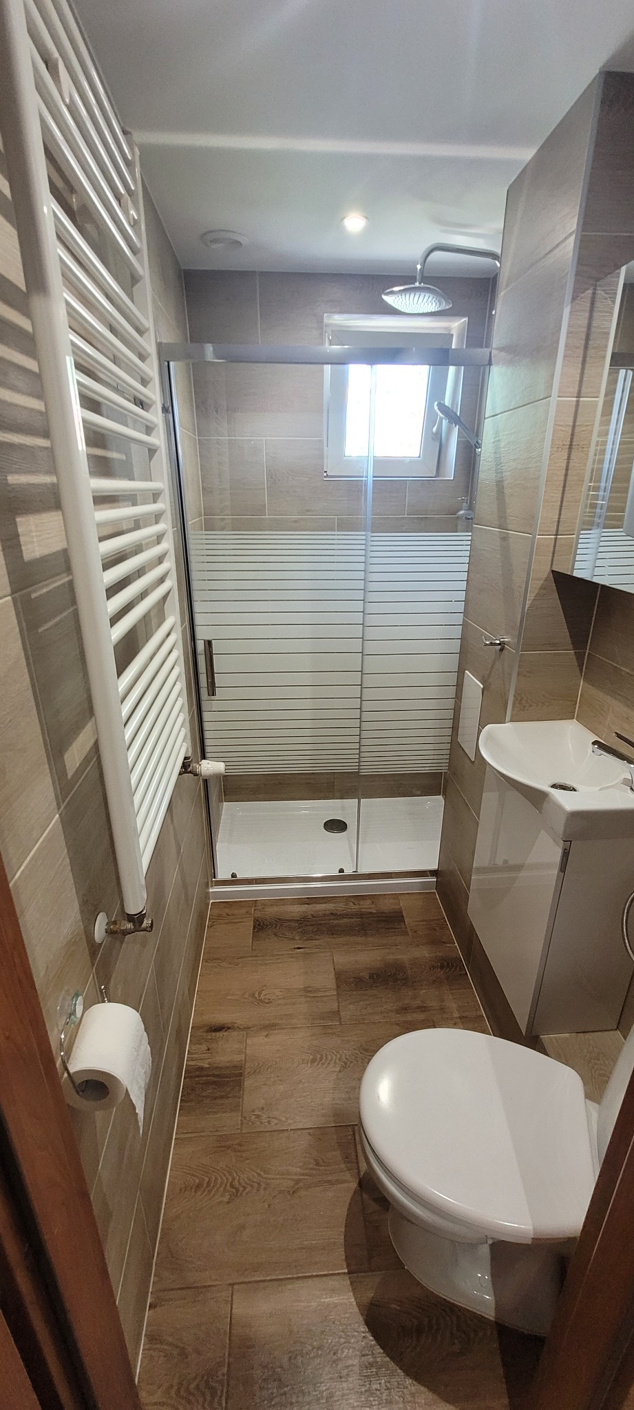 Wąska łazienka po remoncie z kabiną prysznicową z mleczną szybą, grzejnikiem drabinkowym, umywalką z szafką i toaletą, wykończona płytkami w odcieniach brązu i drewnopodobnymi panelami na podłodze.