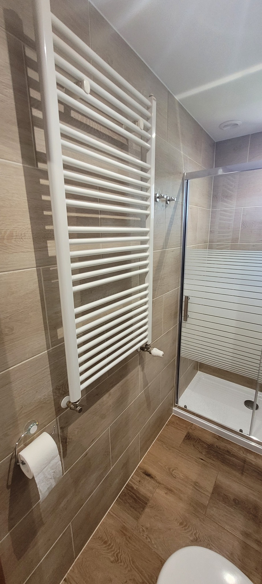 Biały grzejnik drabinkowy na ścianie z płytek imitujących drewno, obok kabina prysznicowa z matową szybą i drewnopodobna podłoga w łazience.