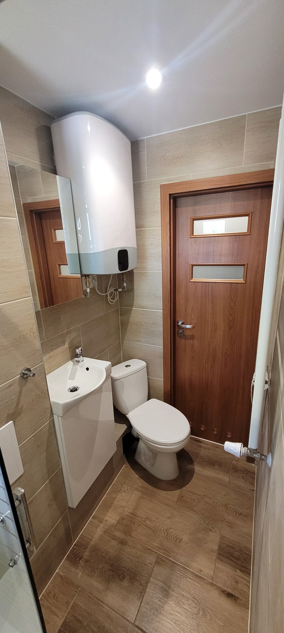 Ukończona łazienka z beżowymi płytkami imitującymi drewno, wiszącą umywalką, toaletą i podgrzewaczem wody nad nią, drzwi z szybkami w tle.