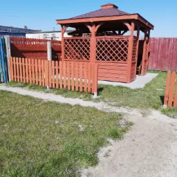 Drewniana altana z kratownicowymi ścianami i brązowym dachem, otoczona niskim płotem z palików, na trawniku w słoneczny dzień.