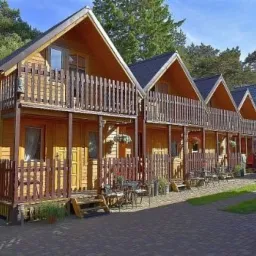 Rząd drewnianych domków letniskowych z balkonami, otoczonych zielenią, w słoneczny dzień. Widoczne meble ogrodowe i fragment zaparkowanego samochodu.