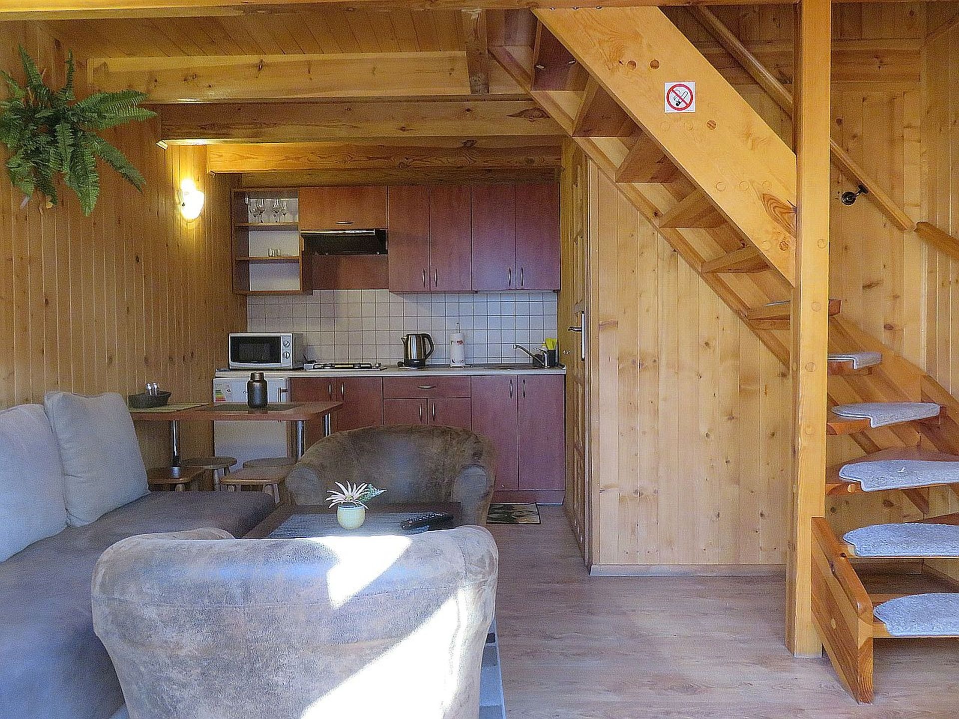 Przytulne wnętrze drewnianego domku letniskowego z aneksem kuchennym, schodami na antresolę i wypoczynkową częścią salonu z sofą.