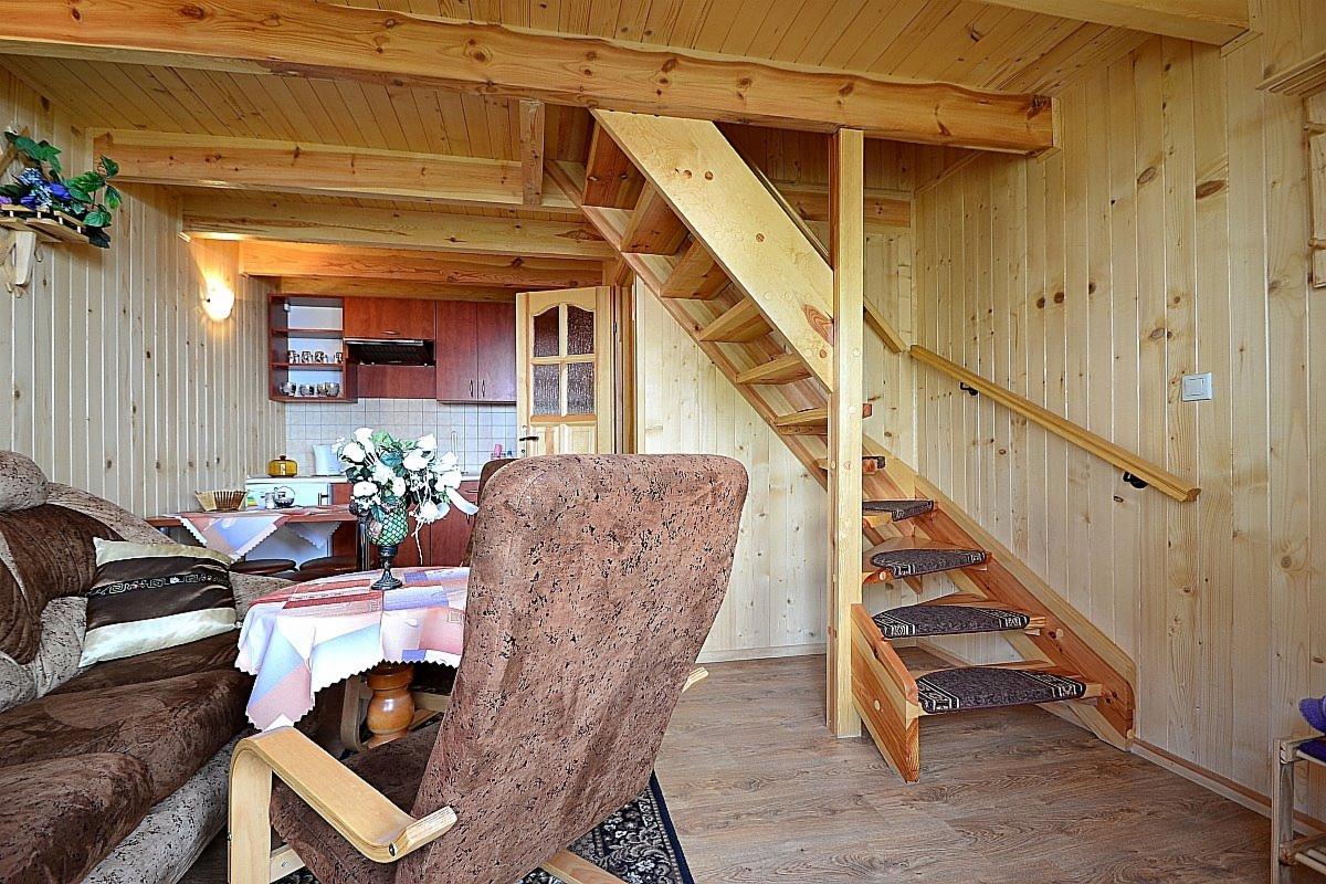 Wnętrze drewnianego domku letniskowego z widokiem na schody, salon z sofą i kuchnię w tle.