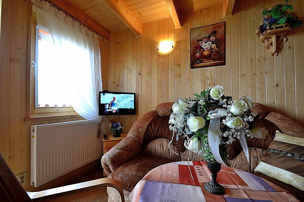 Pokój w drewnianym domku z telewizorem, brązową sofą, bukietem białych róż na stoliku i obrazem kwiatów na ścianie.