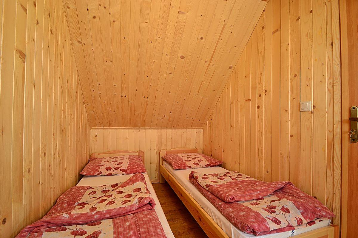 Drewniany pokój z dwoma pojedynczymi łóżkami pod skośnym dachem wykończonym boazerią, czerwono-biała pościel z motywem liści, widoczny fragment drzwi z klamką po prawej stronie.