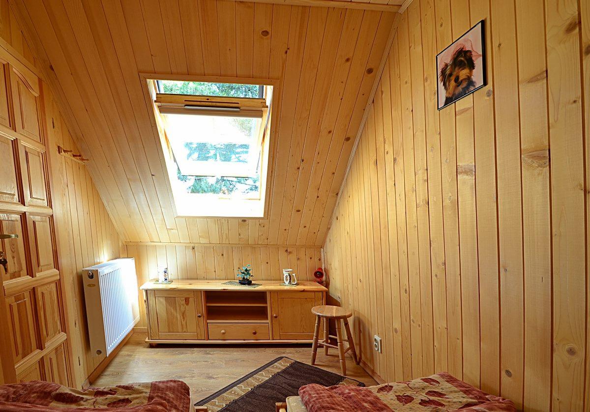 Drewniany pokój na poddaszu z oknem dachowym, meblami z jasnego drewna i dekoracjami świątecznymi na komodzie.