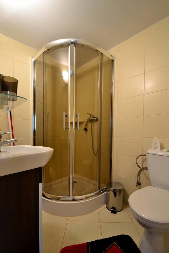 Jasna łazienka z kabiną prysznicową z zaokrąglonymi drzwiami, umywalką z ciemnobrązową szafką, toaletą i metalowym koszem na śmieci; beżowe płytki na ścianach i podłodze.