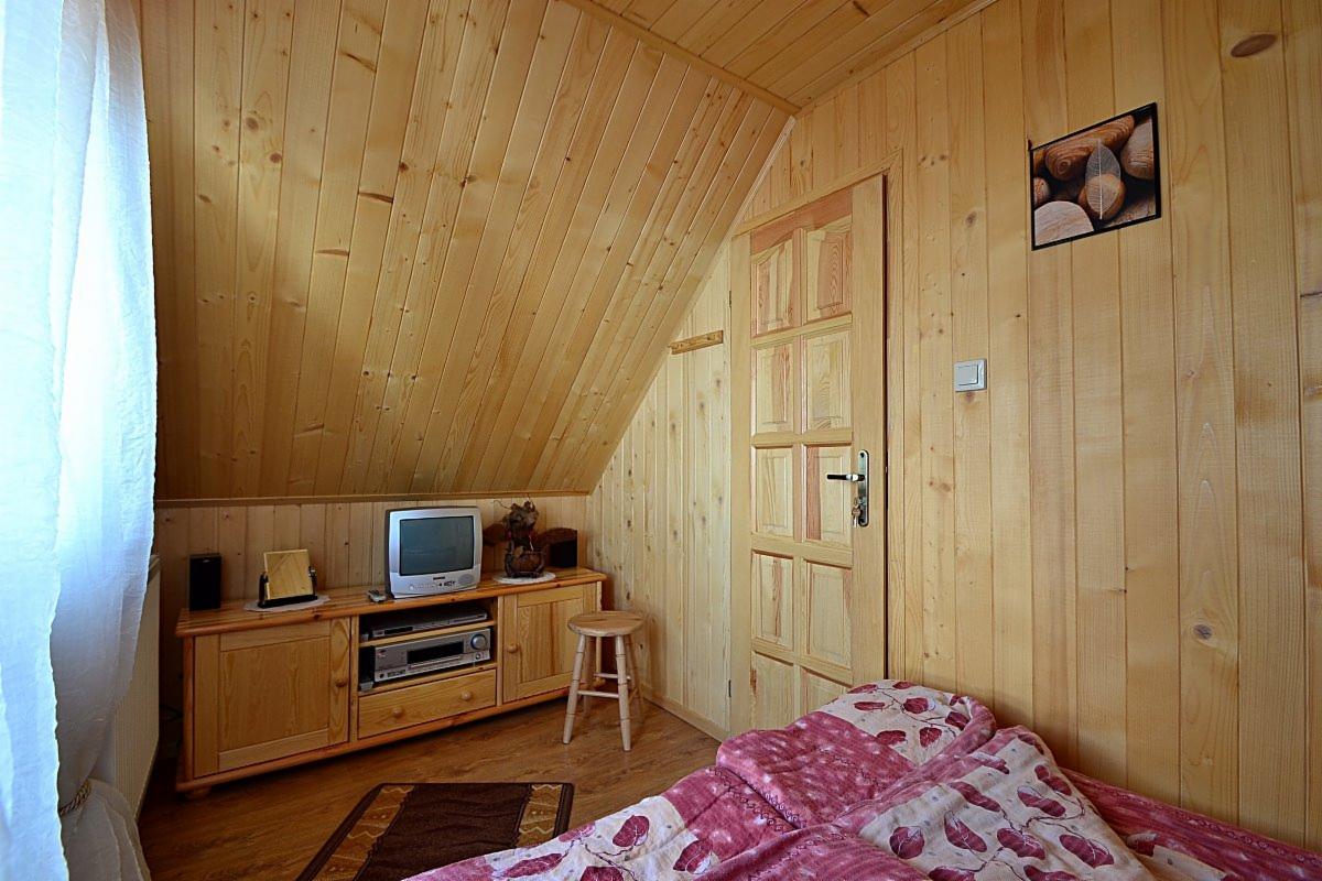 Drewniany pokój na poddaszu z telewizorem, komodą, małym stołkiem i łóżkiem z wzorzystą pościelą; ściany i sufit wyłożone boazerią.
