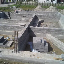 Wykop pod budowę z wylewką fundamentową i częściowo wymurowanymi ścianami z bloczków betonowych, widoczne wystające pręty zbrojeniowe.