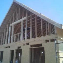 Wznoszenie konstrukcji drewnianej domu z płyt OSB, widoczne rusztowanie i elementy więźby dachowej na tle błękitnego nieba.