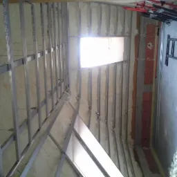 Wnętrze poddasza w trakcie ocieplania pianką PUR, widoczne metalowe profile konstrukcyjne, fragment okna dachowego i narzędzia na podłodze.