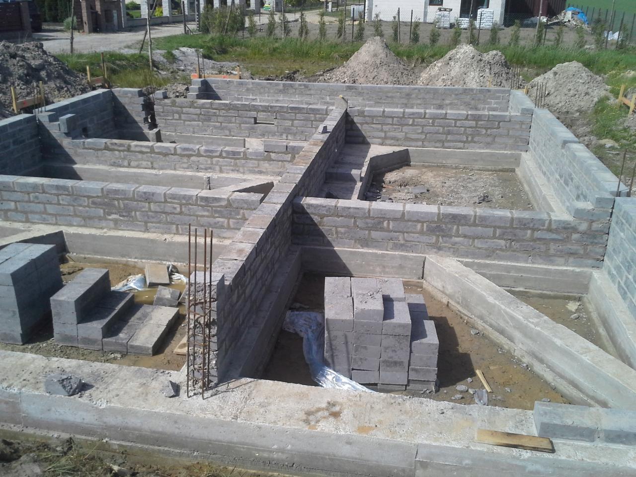 Wykop pod budowę z wylewką fundamentową i częściowo wymurowanymi ścianami z bloczków betonowych, widoczne wystające pręty zbrojeniowe.