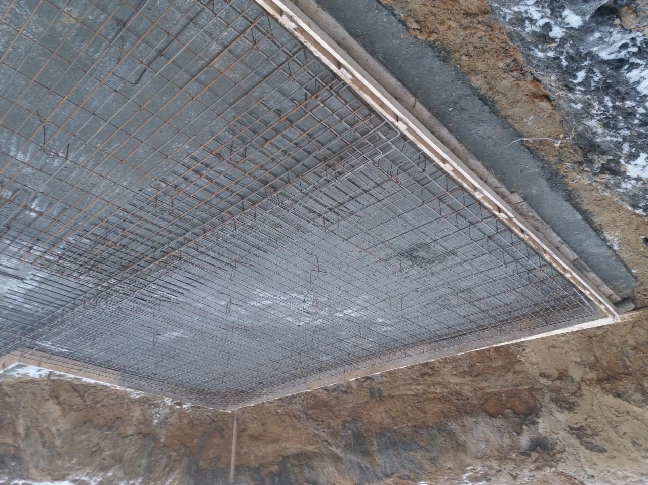 Zbrojenie fundamentu z siatki stalowej, przygotowane do zalania betonem, widoczne drewniane szalunki i wykop ziemny z resztkami śniegu.