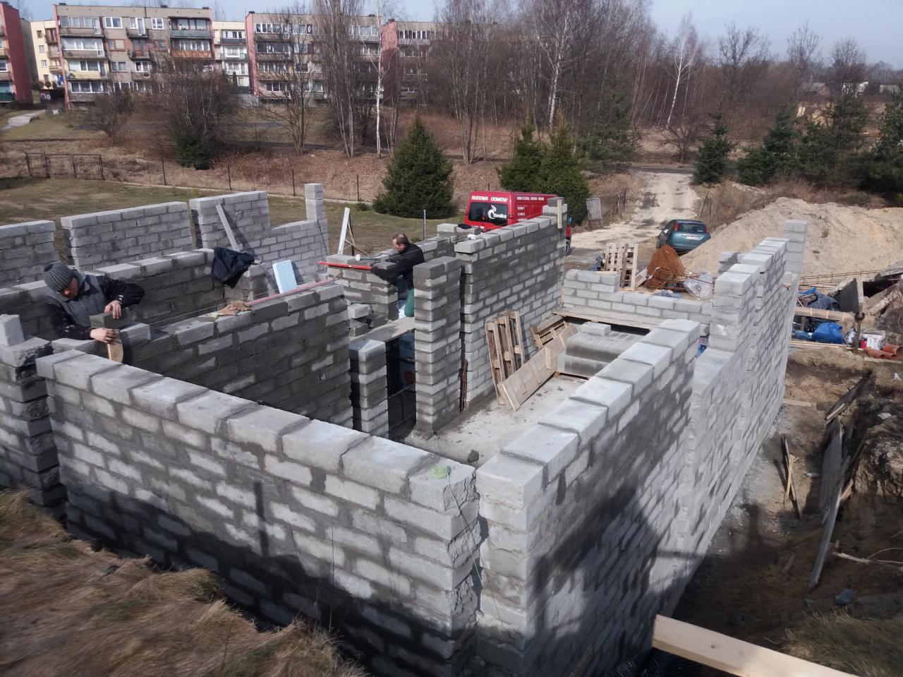 Wznoszenie ścian fundamentowych z bloczków betonowych przez dwóch pracowników, widoczny częściowo wykop i zaparkowany samochód dostawczy na działce budowlanej.