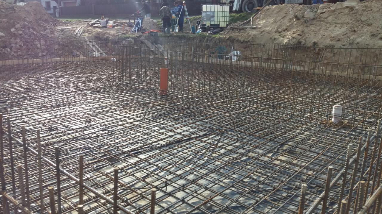 Zbrojenie fundamentu domu jednorodzinnego na folii izolacyjnej, widoczne pionowe rury kanalizacyjne i ekipa budowlana w oddali.