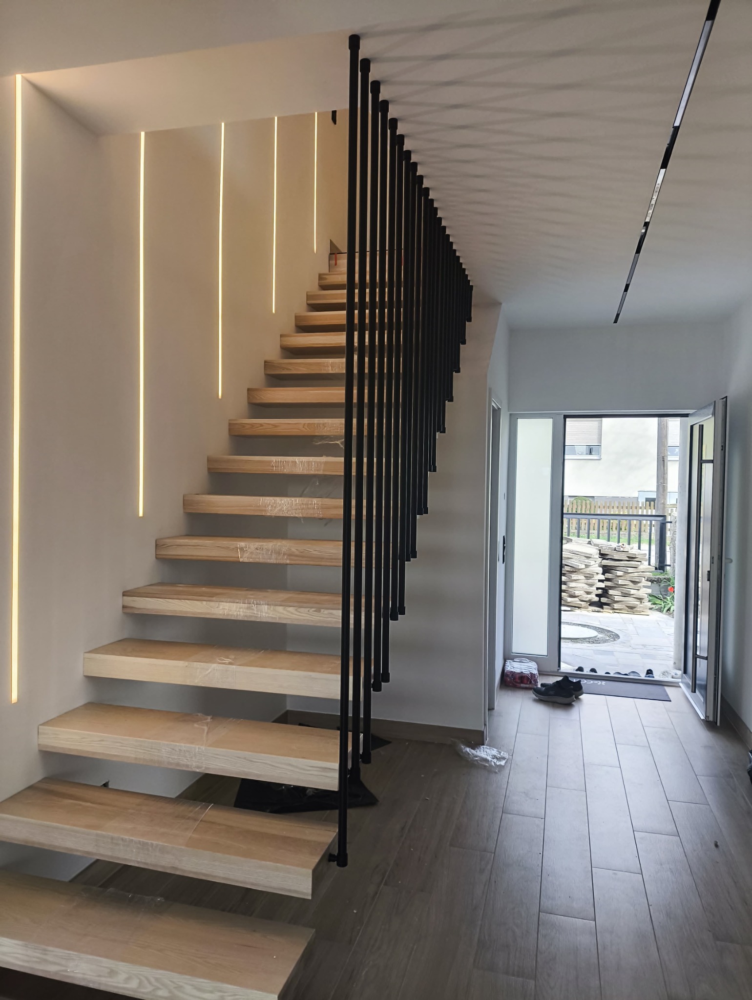 Nowoczesne schody drewniane z czarną balustradą i subtelnym oświetleniem LED wzdłuż ściany, widziane z dołu. Stopnie zabezpieczone folią ochronną.
