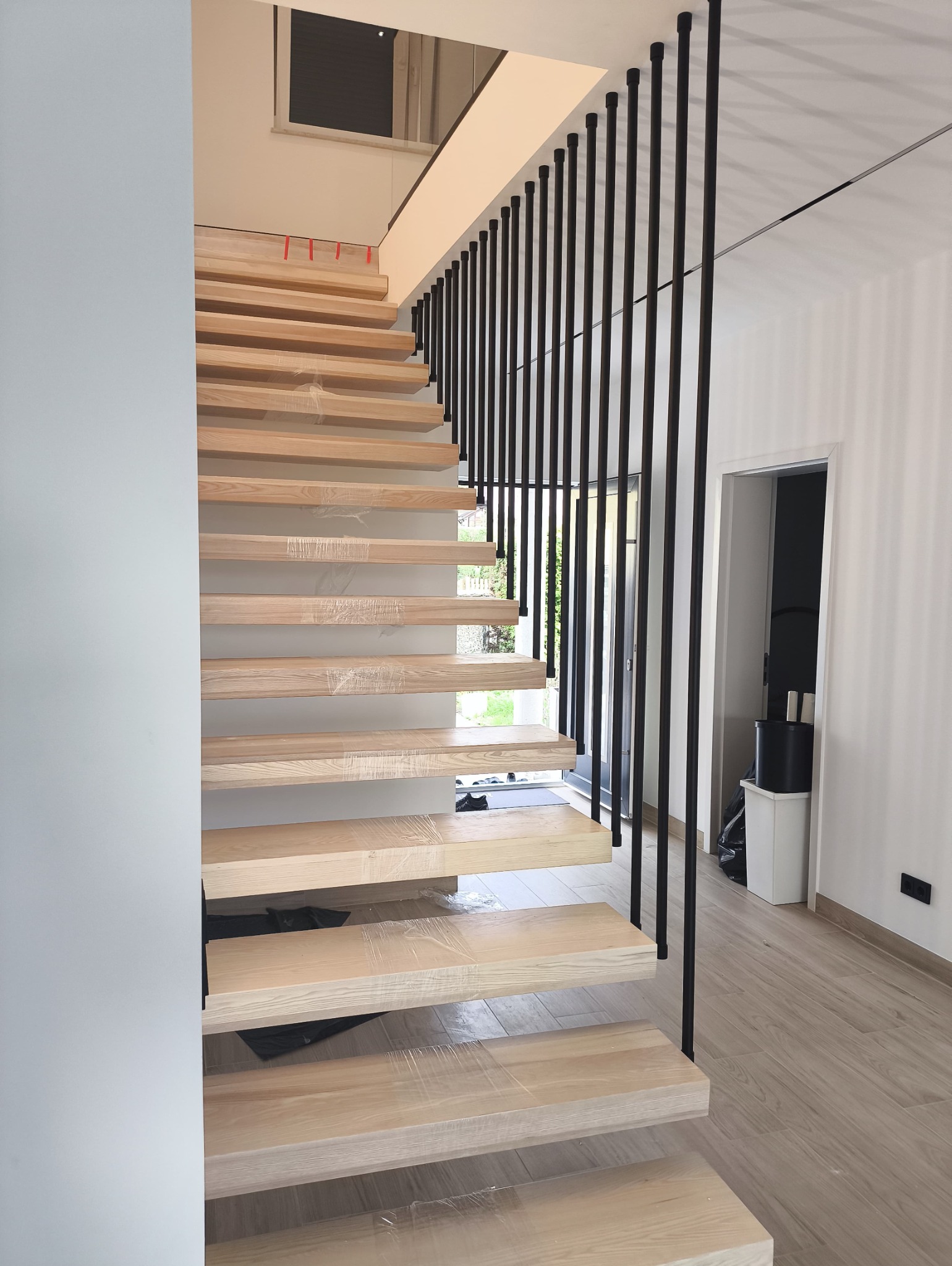 Nowoczesne schody drewniane z czarną balustradą w jasnym wnętrzu. Stopnie zabezpieczone folią ochronną, widoczne elementy wykończenia wnętrza.