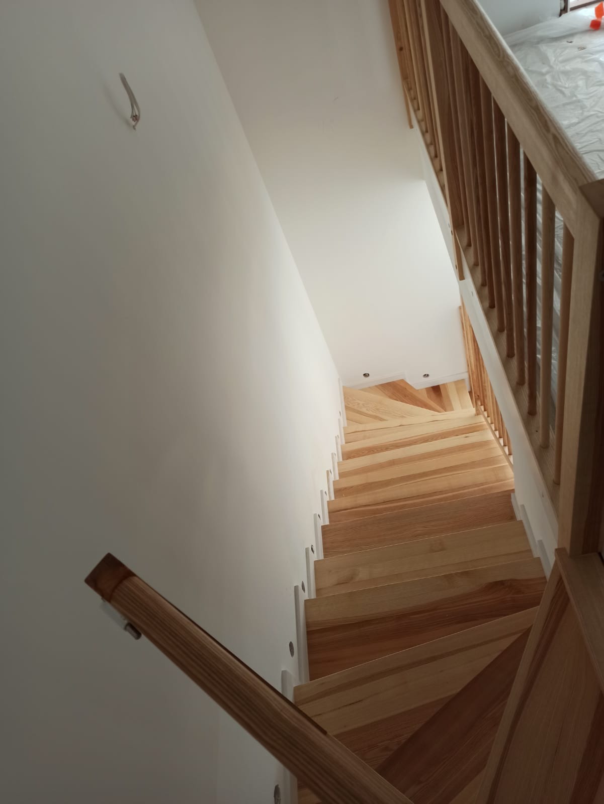 Widok z góry na nowo zamontowane drewniane schody z jasnego drewna z białą balustradą, widoczny fragment białej ściany z niedoskonałościami i wystającym kablem.