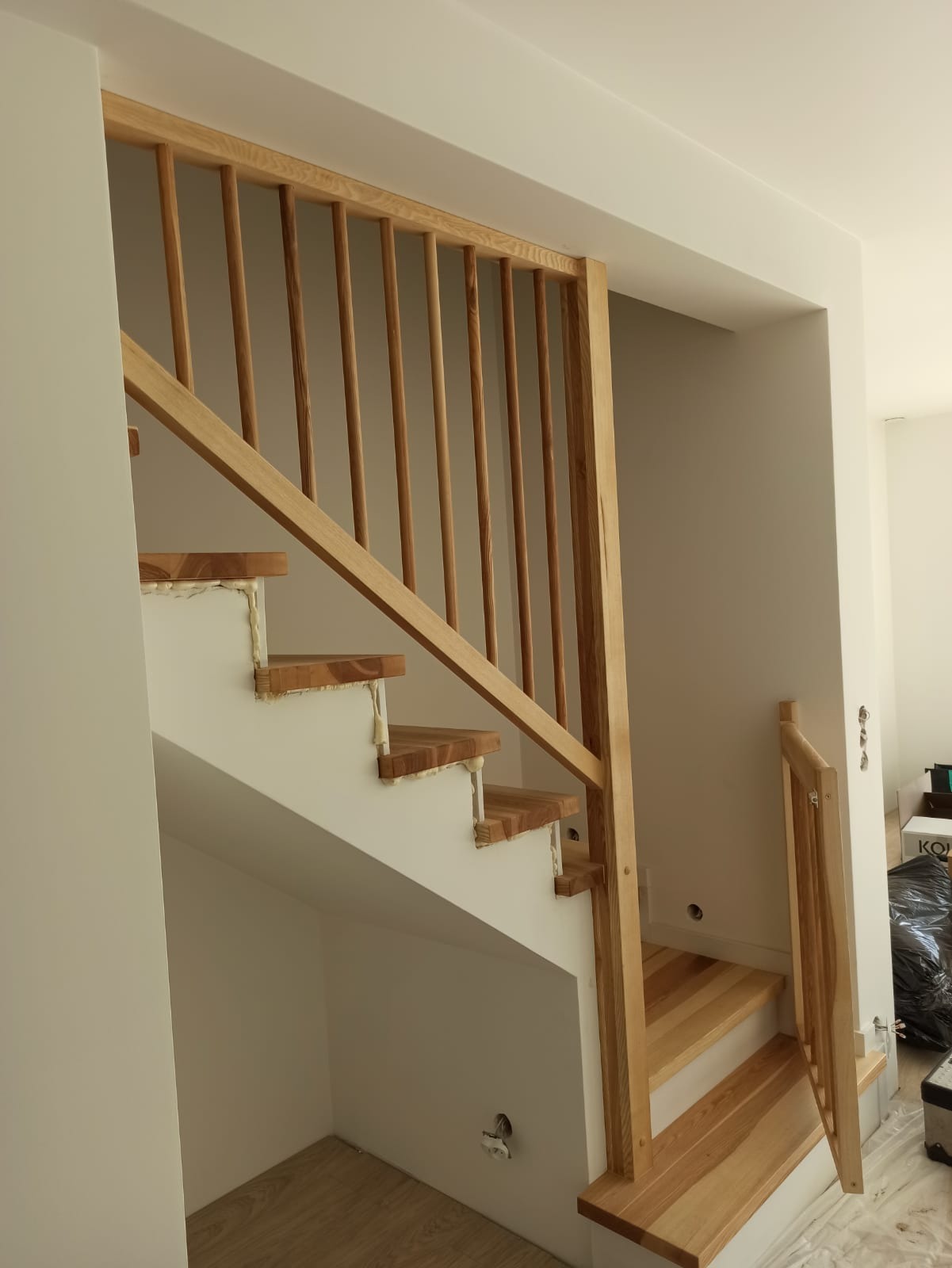 Jasne drewniane schody z balustradą w trakcie montażu, widoczna piana montażowa przy stopniach, minimalistyczne wnętrze w odcieniach bieli i beżu.