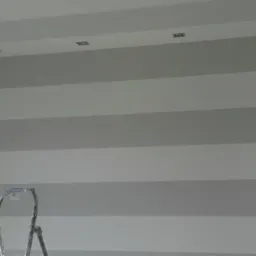 Ściana w pokoju pomalowana w poziome pasy szarości i bieli, widoczna drabina malarska i oświetlenie sufitowe