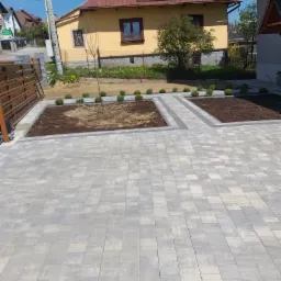 Szara kostka brukowa ułożona na podjeździe z przygotowanymi rabatami i młodymi drzewkami w tle żółtego, dwupiętrowego domu.