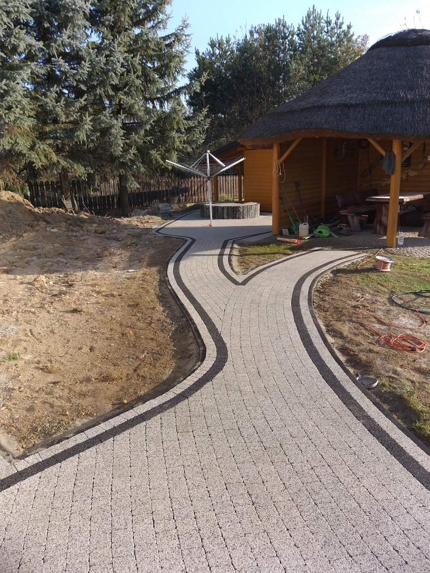 Ułożona ścieżka z kostki brukowej w ogrodzie, z czarnymi obrzeżami, prowadząca do altany z trzcinowym dachem i suszarki na pranie.