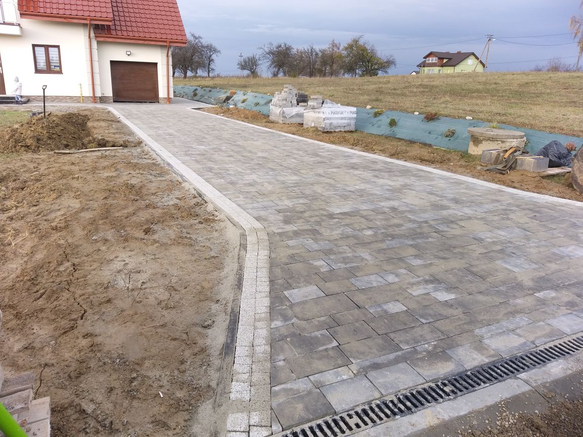 Ułożona, szara kostka brukowa z jasną obwódką prowadząca od domu z brązową bramą garażową, widoczny drenaż liniowy, teren częściowo przygotowany pod dalsze prace.