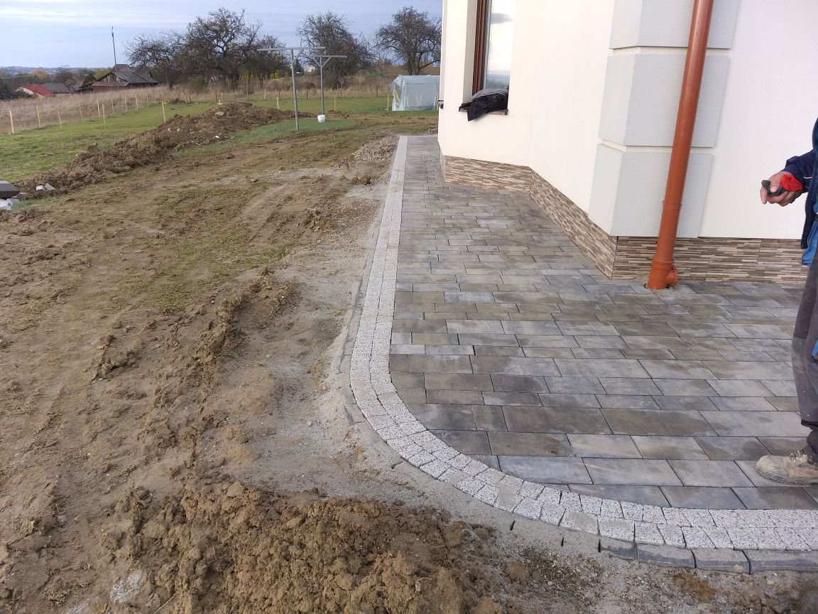 Szara kostka brukowa ułożona przy ścianie budynku z brązową rynną, obrzeże z drobnej kostki, obok teren przygotowany pod dalsze prace ziemne.