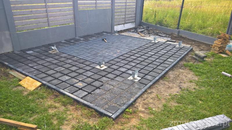 Częściowo ułożona, kwadratowa, ciemnoszara kostka brukowa w metalowej ramie, widoczne metalowe wsporniki, młotek oraz stos kostki brukowej i fragment ogrodzenia panelowego w tle.