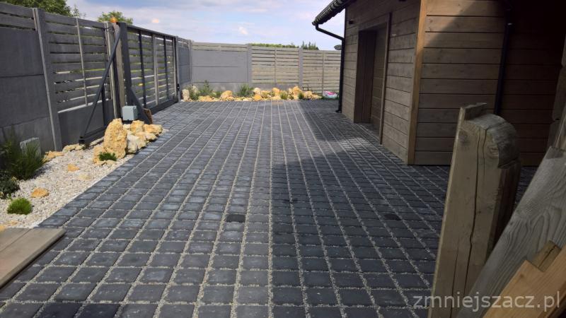 Ułożona, ciemnoszara kostka brukowa z jasnymi fugami, otoczona szarym ogrodzeniem panelowym i drewnianym budynkiem gospodarczym, z elementami dekoracyjnymi z kamieni i roślinności.