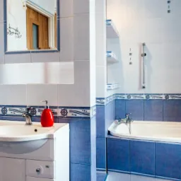 Wykończona łazienka z niebiesko-białymi płytkami, białą szafką pod umywalką, wanną i akcesoriami łazienkowymi, takimi jak czerwony dozownik do mydła.