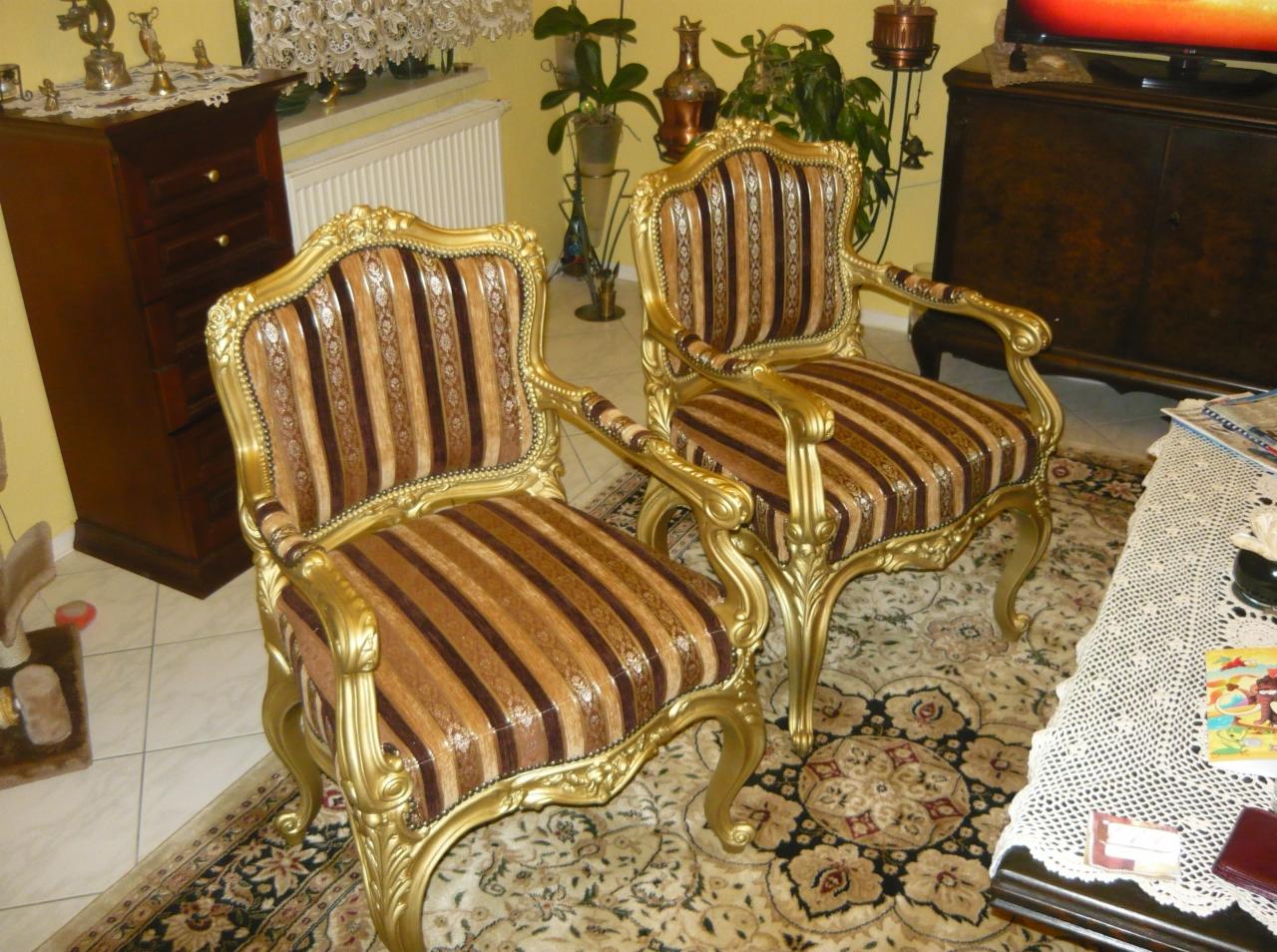 Dwa bogato zdobione, złote fotele z tapicerką w brązowe pasy, stojące na wzorzystym dywanie w salonie z ciemnymi meblami w tle.