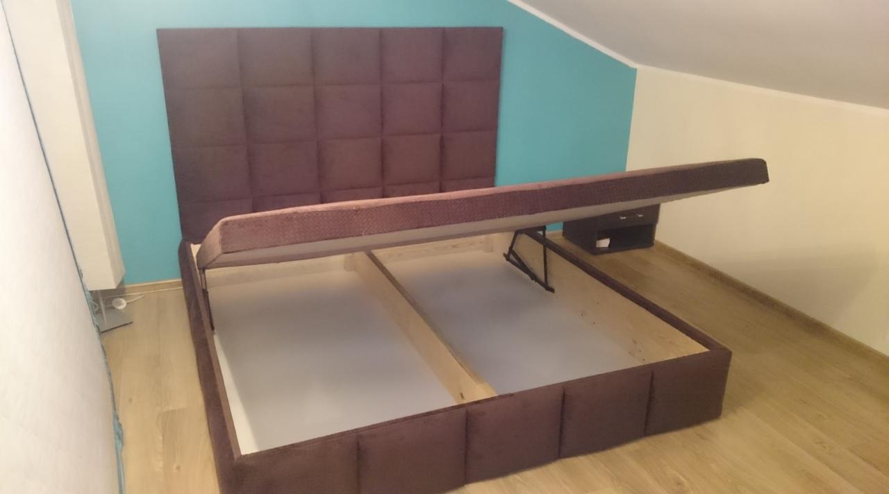 Rozłożone łóżko tapicerowane z brązowym wezgłowiem w kwadraty i podnoszonym stelażem, widoczna pusta przestrzeń do przechowywania, na podłodze panele.