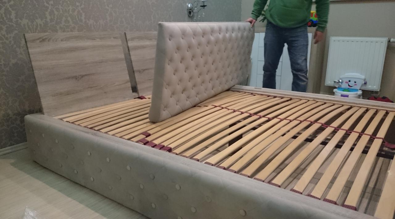Montaż tapicerowanego łóżka z drewnianym zagłówkiem i stelażem z listew, widoczny fragment osoby w tle.