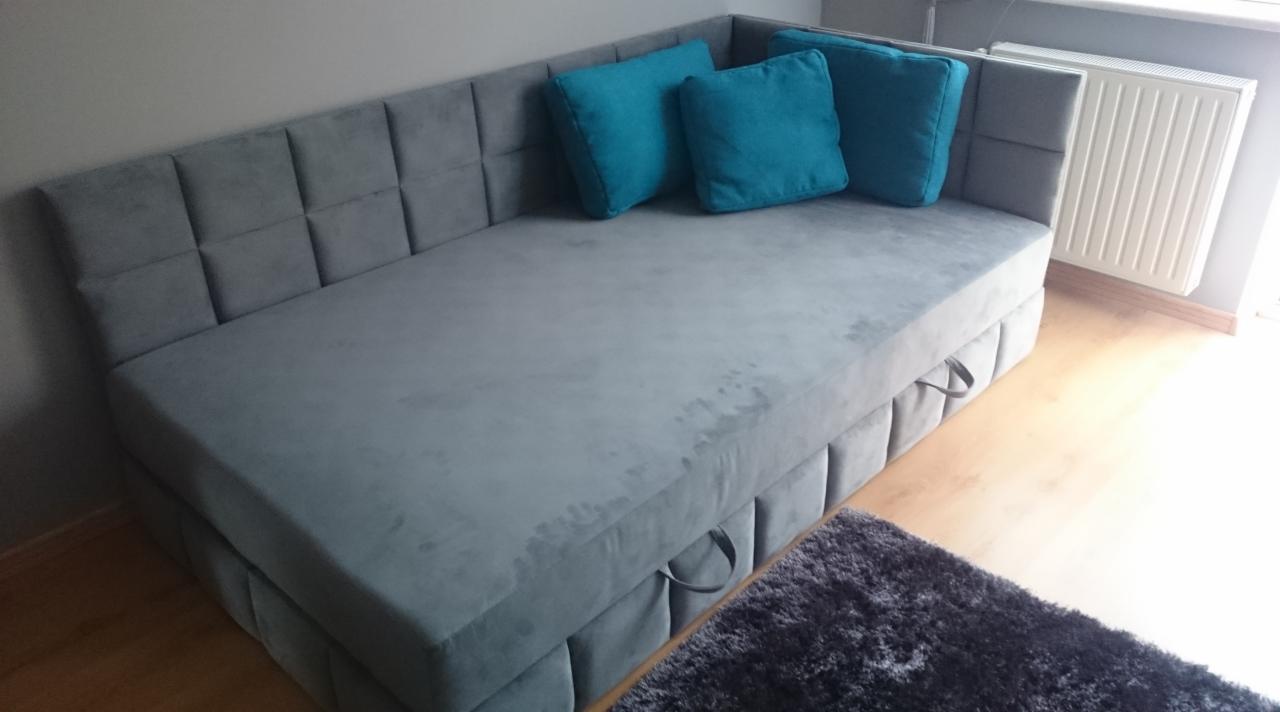 Szara, tapicerowana sofa z pikowanym wezgłowiem i turkusowymi poduszkami, widoczny fragment dywanu i grzejnik.