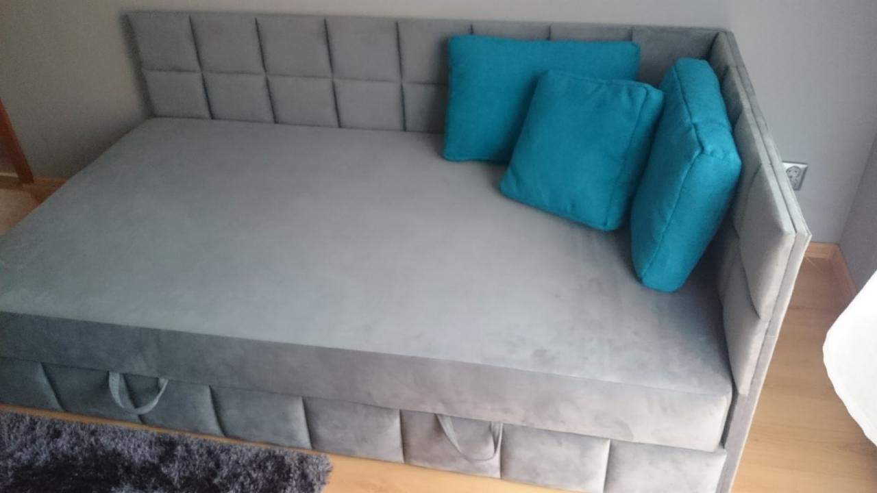 Szary tapicerowany narożnik z pikowanym zagłówkiem i turkusowymi poduszkami, widoczne uchwyty szuflad pod siedziskiem.