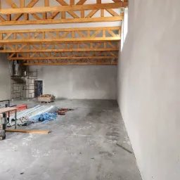 Wnętrze budynku w trakcie tynkowania maszynowego; widoczny pracownik na rusztowaniu, świeżo nałożony tynk na ścianach oraz drewniana konstrukcja stropu.