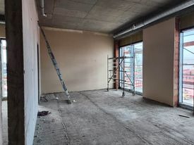 Surowe wnętrze w trakcie prac wykończeniowych: ściany pokryte świeżym, jasnym tynkiem, widoczne rusztowanie i drabina oparta o ścianę, surowy betonowy sufit z instalacjami, okna z widokiem na budowę.