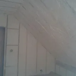 Poddasze z widoczną izolacją natryskową z piany, częściowo zakryte drewniane elementy konstrukcyjne, pionowe listwy montażowe i zamknięty otwór drzwiowy z płyty OSB.
