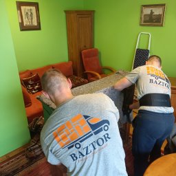 Baztor Transport Przeprowadzki - Dwóch mężczyzn w szarych koszulkach z logo firmy transportowej Baztor przenosi mebel owinięty w szary materiał w zielonym pokoju z dywanem o wzorzystym deseniu.