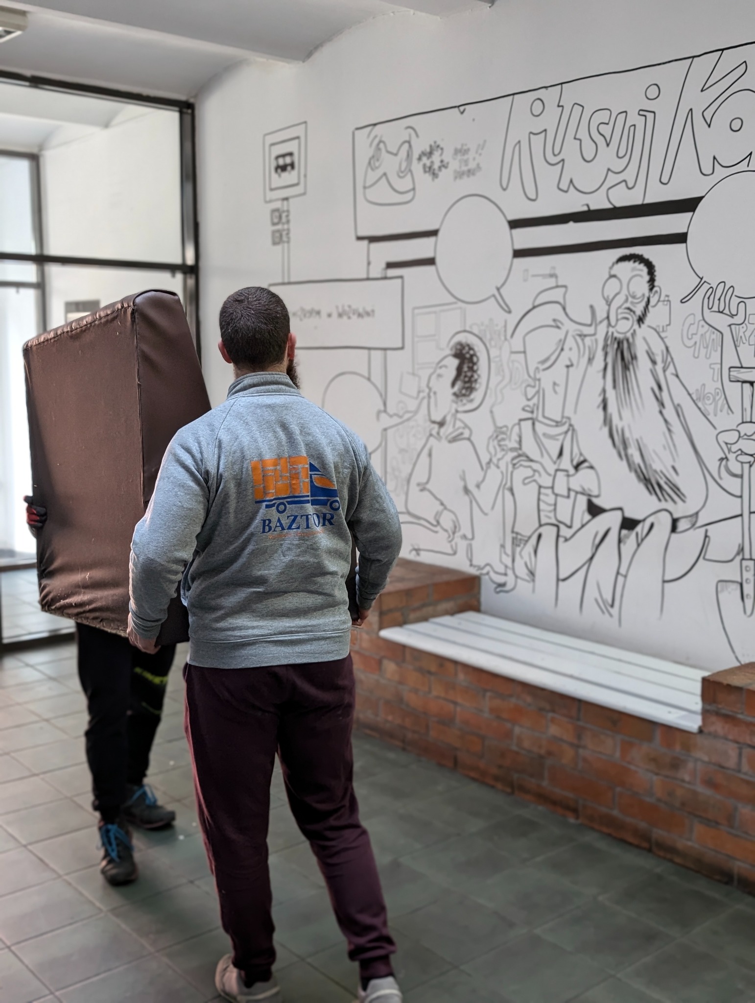 Dwóch mężczyzn przenosi brązowy mebel tapicerowany w korytarzu z rysunkami na ścianie; widoczny tył bluzy z logo firmy transportowej 'BAZTOR'.